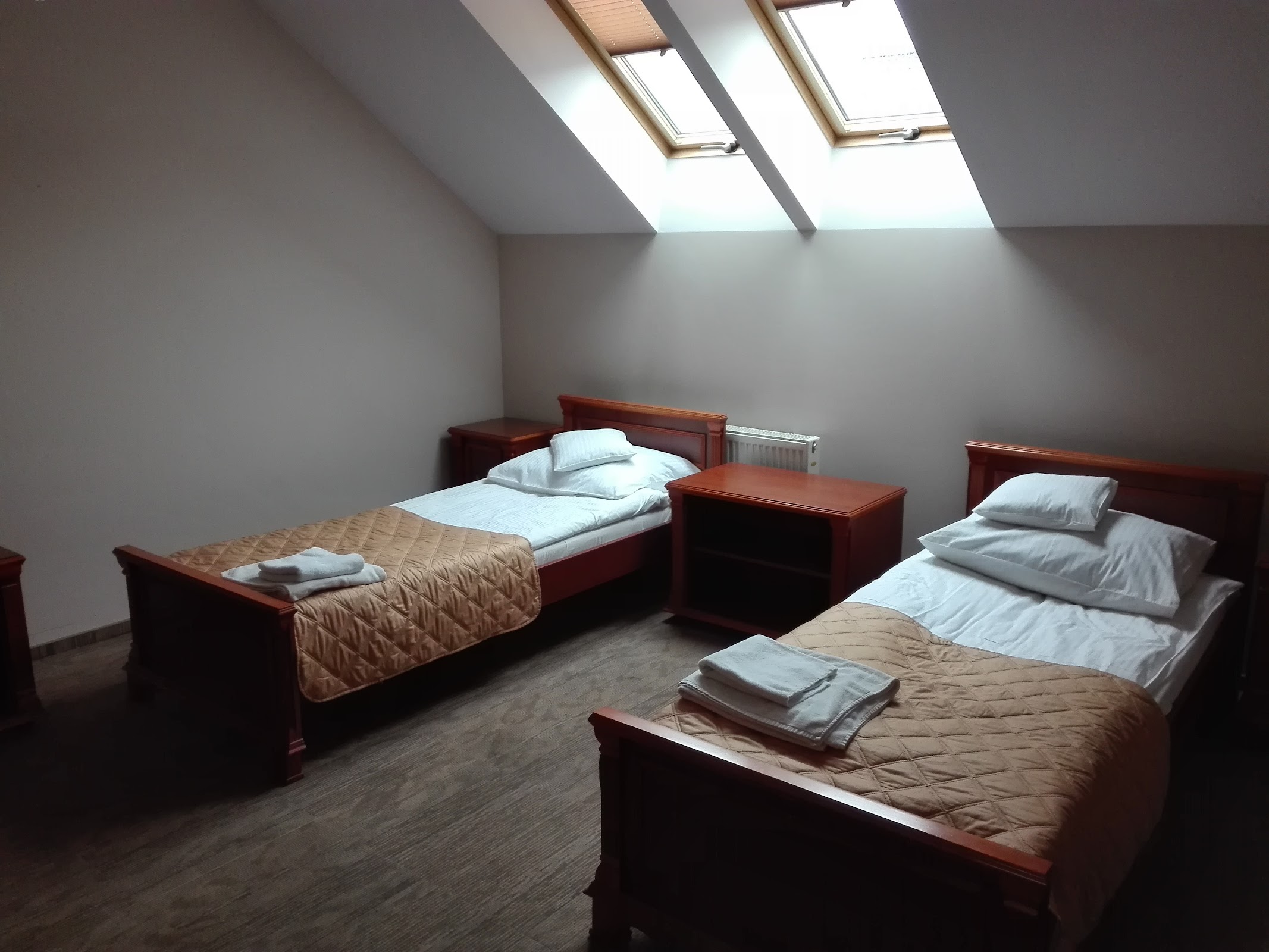 Pokój dwuosobowy na poddaszu Hotelu Biesiada - dwa łóżka pojedyncze z mahoniowymi ramami, beżowe narzuty, drewniana szafka nocna, okna dachowe, ciepłe oświetlenie
