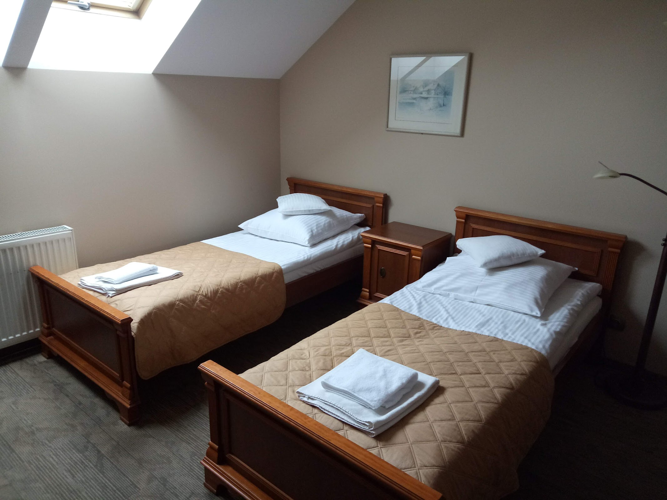 Pokój dwuosobowy w Hotelu Biesiada - dwa łóżka z drewnianymi zagłówkami, kremowe ściany, akwarela na ścianie, okno dachowe, lampa stojąca, dywanowa wykładzina