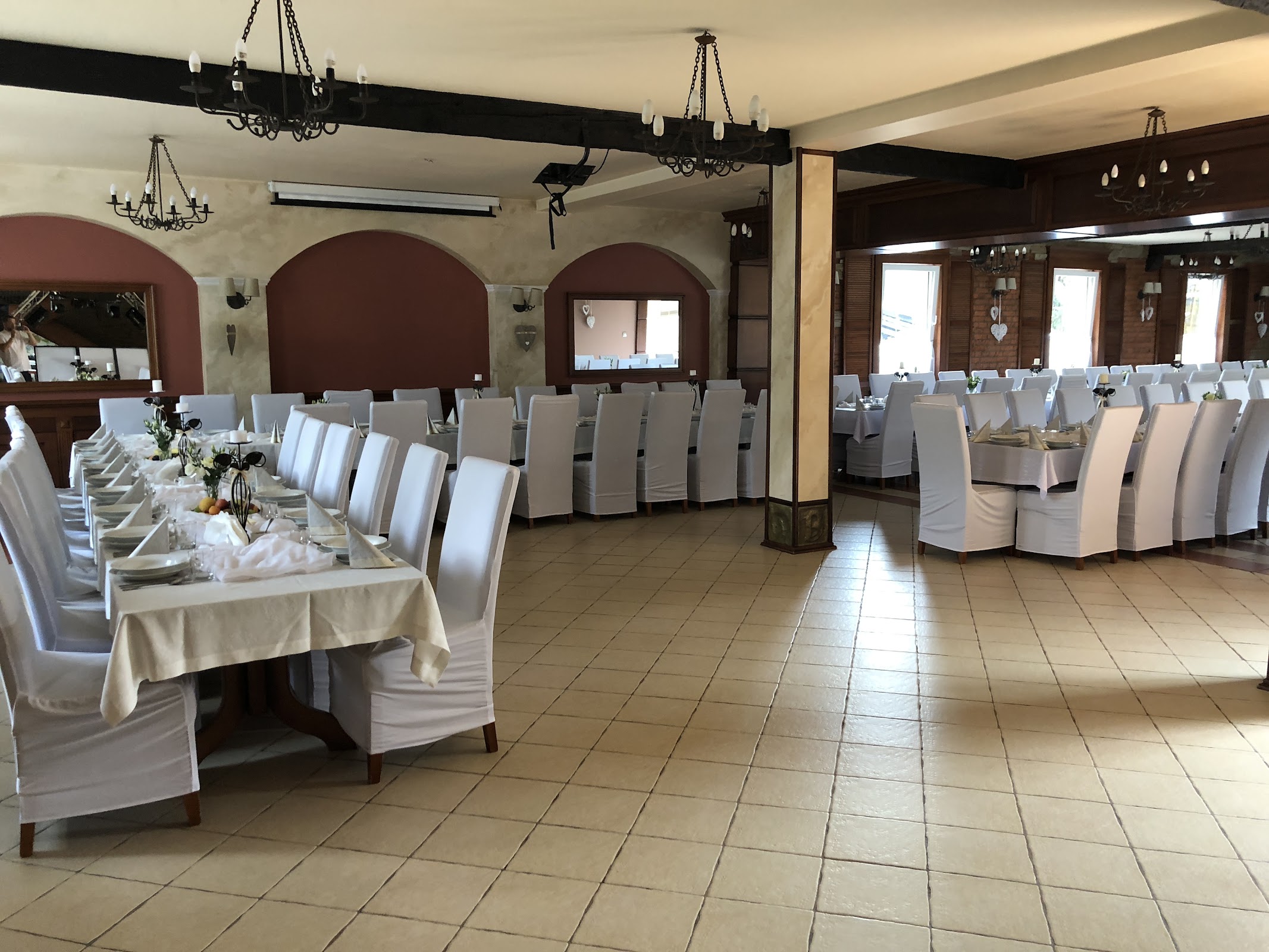 Sala bankietowa Restauracji Biesiada - długi stół na 50 osób nakryty białym obrusem, krzesła w białych pokrowcach, kute żyrandole, łukowe okna z bordowym wykończeniem