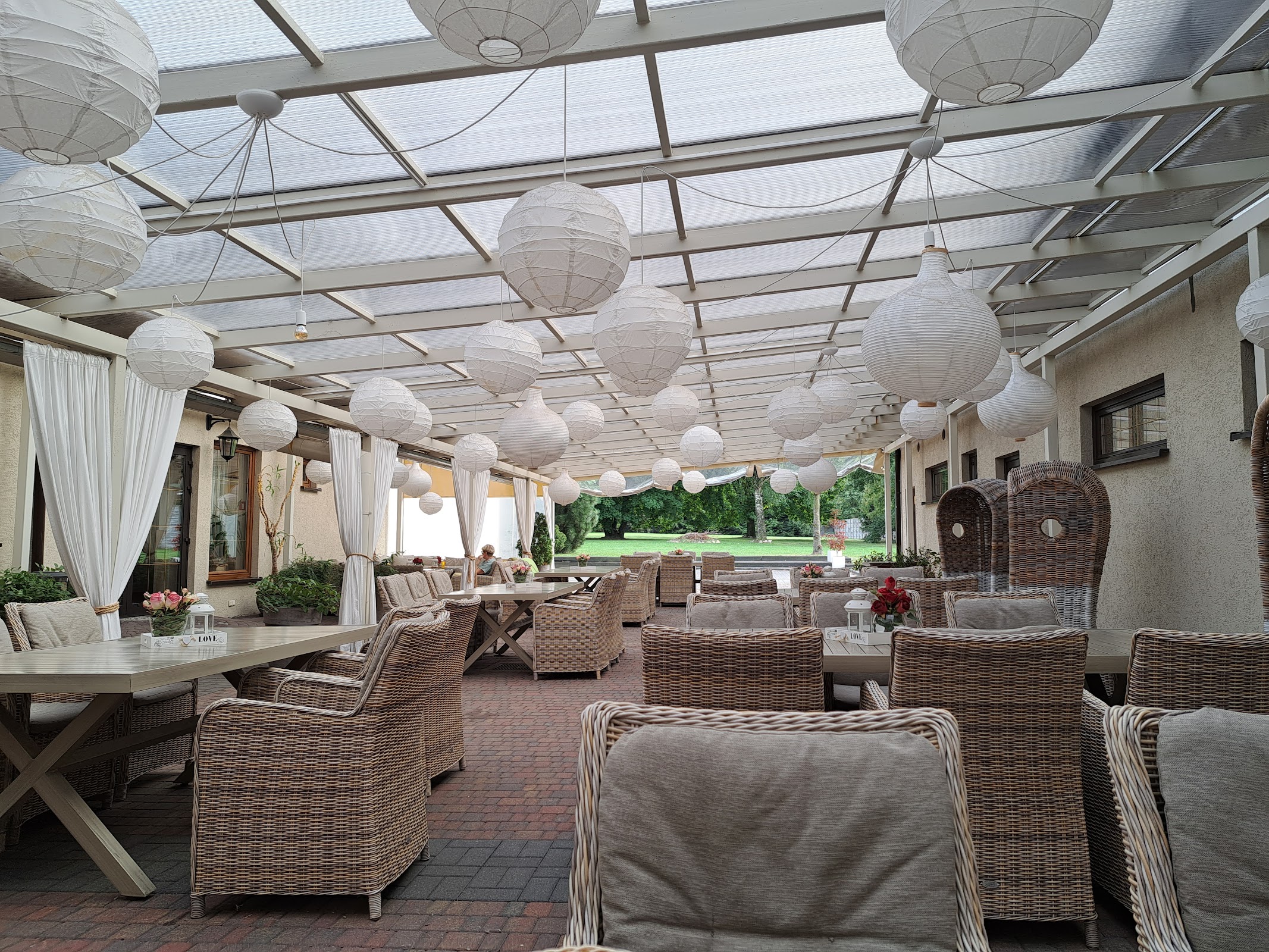 Patio restauracyjne Hotelu Biesiada - zadaszona przestrzeń, wiklinowe fotele, lniane białe kotary, papierowe lampiony, długie stoły z naturalnego drewna, widok na ogród z trawnikiem